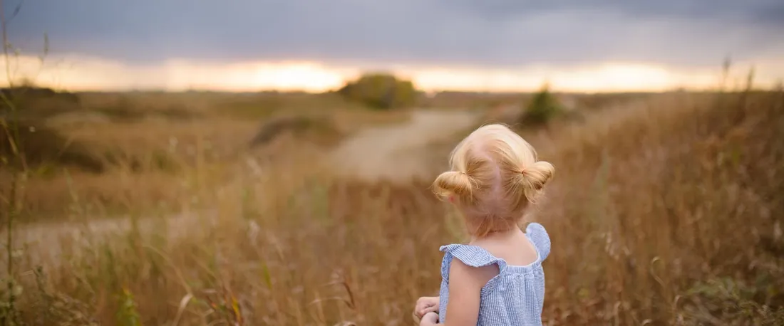 Une petite fille de dos dans un champ doré, qui regarde un ciel d'orage (photo d'ambiance)